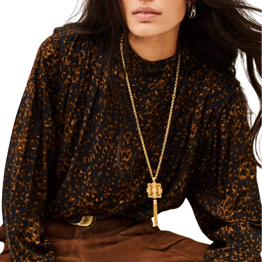 A woman wearing a black and brown leopard-print long-sleeve top, brown pants, and a long gold pendant necklace, seated against a plain white background.